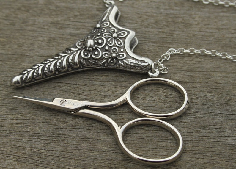 May include: A silver necklace with a floral design pendant and a pair of small silver scissors. The pendant is shaped like a teardrop and has a chain attached to it. The scissors are closed and have a silver finish.