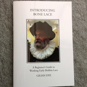 May include: A white book cover with the title "Introducing Bone Lace" and the subtitle "A Beginner's Guide to Working Early Bobbin Lace" by Gillian Dye. A black and white photo of a person wearing a ruff and a hat is on the cover.