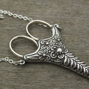 May include: A silver necklace with a pendant shaped like a pair of scissors with a floral design. The pendant is detailed with leaves and flowers.