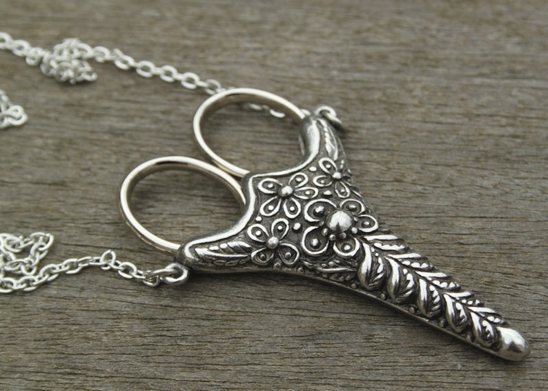 May include: A silver necklace with a pendant shaped like a pair of scissors with a floral design. The pendant is detailed with leaves and flowers.