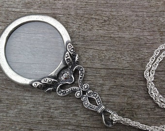 May include: A silver-toned magnifying glass pendant necklace with an ornate handle and a decorative frame. The round lens is set in a detailed bezel, and the pendant hangs from a delicate chain. The design features floral and scrollwork elements.