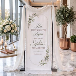 May include: A vertical white fabric banner hanging on a black metal stand, draped with sheer white fabric. The banner features an arched border, green leaf illustrations, and the text: Her NEXT CHAPTER Begins WELCOME TO Sophia's BRIDAL SHOWER MAY 24, 2026.