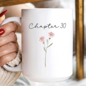 Puede incluir: Taza de cerámica blanca con el texto "Chapter 30" en escritura negra y un diseño floral en acuarela. El diseño floral presenta dos flores rosas con tallos y hojas verdes. La taza tiene un asa grande.