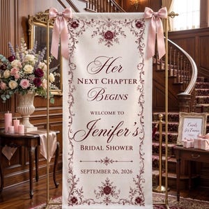 May include: A vertical cream-colored fabric banner featuring dark red floral scrollwork borders and two dusty pink satin bows at the top. The text reads: Her NEXT CHAPTER BEGINS WELCOME TO Jenifer's BRIDAL SHOWER SEPTEMBER 26, 2026, displayed on a gold stand.