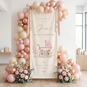 Puede incluir: Un cartel vertical de color crema para un baby shower con una ilustración de un cochecito vintage rosa, detalles florales y el texto "Welcome to Jean's Baby Shower" y "oh baby, we can't wait to meet you," enmarcado por un arco de globos en rosa y dorado.