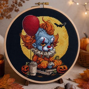 May include: A circular wooden embroidery hoop containing a cross-stitch design of a clown with orange hair, a red balloon, and a coffee cup against a yellow moon and black background, surrounded by small orange pumpkins and a flying bat silhouette.