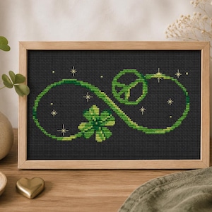 May include: A framed cross-stitch embroidery on black fabric featuring a bright green infinity symbol. The design incorporates a four-leaf clover, a peace sign, and small yellow star accents. The piece is enclosed in a light-toned wooden frame.