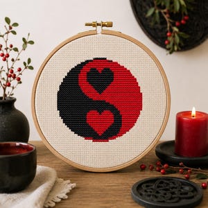 Puede incluir: Un bastidor de bordado circular de madera que contiene un diseño de punto de cruz de un símbolo yin-yang. El símbolo se compone de una sección negra con un corazón rojo y una sección roja con un corazón negro, bordado sobre tela beige.