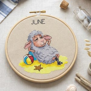 Può includere: Un telaio da ricamo circolare in legno contenente un disegno a punto croce su tessuto beige. Il design raffigura una pecora grigia che riposa sulla sabbia gialla con un pallone da spiaggia colorato e una stella marina marrone. Il testo JUNE è ricamato in nero.