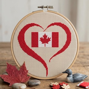 Puede incluir: Un bastidor de bordado circular de madera que contiene un patrón de punto de cruz de un corazón rojo que enmarca la bandera canadiense. El diseño presenta una hoja de arce roja sobre fondo blanco con paneles laterales rojos, junto a una hoja.