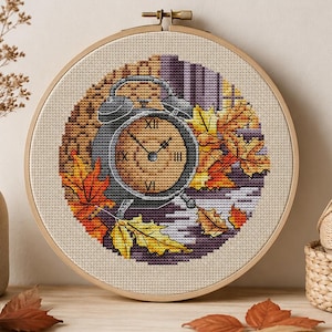 May include: A circular wooden embroidery hoop containing a cross-stitch pattern of a grey alarm clock surrounded by orange and yellow autumn leaves. The clock face features Roman numerals and is set against a background of purple and brown tones.