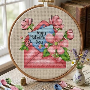 Może przedstawiać: Tamborek do haftu krzyżykowego z różowym wzorem koperty, kwiatami i napisem "Happy Mother's Day" na niebieskiej karcie. Scena obejmuje nici do haftu, nożyczki i mały wazon z kwiatami.