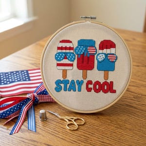 May include: A cross-stitch embroidery hoop featuring three popsicle designs with sunglasses, in red, white, and blue. The text "STAY COOL" is stitched below the popsicles. An American flag and scissors are in the background.