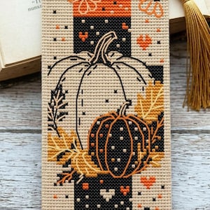 Può includere: Un segnalibro rettangolare a punto croce su tela Aida beige con il contorno di una grande zucca bianca e una zucca nera più piccola con pois arancioni. Il design include fiori, foglie e cuori arancioni su uno sfondo a righe nere e arancioni, rifinito con una nappa dorata.