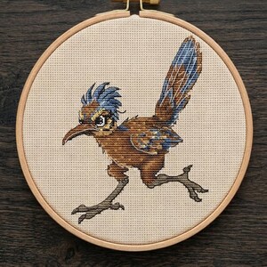 May include: A cross-stitch embroidery of a roadrunner bird in a circular wooden hoop. The bird features brown and blue plumage, a long tail, and a sharp beak, stitched onto a light beige fabric background with a visible grid pattern.