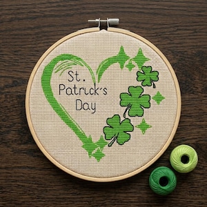 May include: A cross-stitch embroidery hoop with a St. Patrick's Day theme. The design features a green heart, shamrocks, and the text "St. Patrick's Day." Two green spools of thread are next to the hoop.