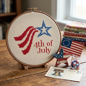May include: A circular wooden embroidery hoop containing a cross-stitch design on cream fabric. The design features a red striped flag motif, a blue star, and the red text 4th of July. The hoop is displayed alongside a small star-shaped ornament.