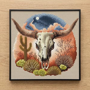 May include: Framed cross-stitch artwork featuring a bull skull with large horns, set against a desert landscape. The design includes a cactus, shrubs, and a night sky with a moon. The color palette includes shades of brown, orange, and blue.