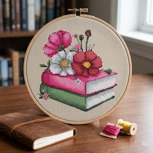 May include: Cross-stitch embroidery in a wooden hoop featuring a stack of books with pink and green covers, topped with pink, white, and red flowers. A brown leather journal and spools of thread are in the foreground.