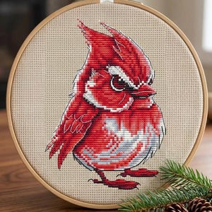 Puede incluir: Un proyecto de bordado en punto de cruz dentro de un bastidor de madera redondo que muestra un pájaro cardenal rojo estilizado con expresión gruñona, detalles blancos y una cresta puntiaguda sobre tela beige claro con cuadrícula.
