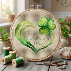Può includere: Un telaio da ricamo a punto croce con un disegno per la festa di San Patrizio. Il disegno presenta un trifoglio verde e una forma di cuore con la scritta "St. Patrick's Day". Il ricamo è ambientato in un telaio di legno, con rocchetti di filo verde e forbici nelle vicinanze.