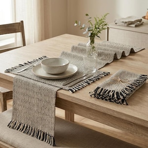 Pode incluir: Uma mesa de jantar com um caminho de mesa tecido bege e preto, individuais e uma toalha de mesa a condizer. Uma tigela e um prato brancos estão colocados sobre o caminho de mesa. Um vaso de vegetação e um copo de água também estão na mesa.