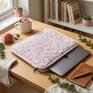 May include: A quilted laptop sleeve featuring a light pink fabric with a green and white floral pattern. The sleeve has a white zip closure and is partially open, revealing a dark grey laptop inside. The design includes a diamond-stitched texture.