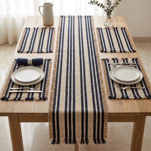 May include: A dining table setting featuring a beige and navy blue striped table runner, placemats, and napkins. White plates, silverware, and a pitcher are also on the table. The table setting has a rustic, farmhouse style.