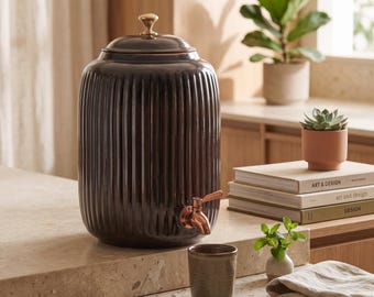 May include: A dark brown, vertically ribbed ceramic water dispenser with a metallic copper-toned spigot and a matching lid topped with a small, round gold-coloured knob. The cylindrical vessel features a glossy finish and a wide, rounded body.