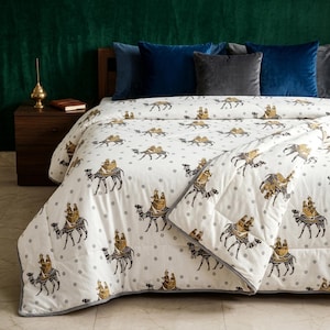 May include: White comforter with a repeating pattern of camels in black and gold. The comforter has a gray border and is placed on a bed with blue and gray pillows. The background is a dark green wall.