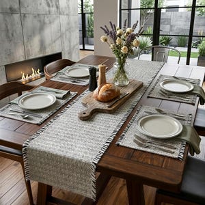 Pode incluir: Uma mesa de jantar de madeira escura com um caminho de mesa de padrão cinza e branco e jogos americanos a condizer. A mesa apresenta pratos de cerâmica branca, talheres de prata, guardanapos verde-azeitona, um vaso de vidro e pão.