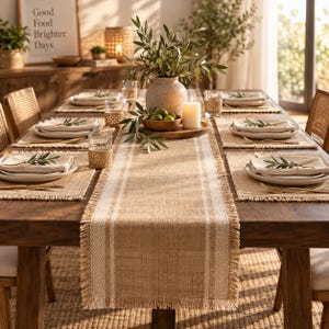 May include: A rectangular burlap table runner with white vertical stripes and fringed edges, centered on a wooden dining table. The runner features a woven texture and is decorated with a ceramic vase, green foliage, and a white pillar candle.