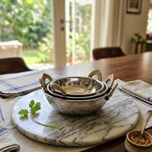 May include: A set of two nested stainless steel bowls with polished interiors and textured gold-toned metal handles. The bowls feature a rounded base and are positioned on a circular white marble board with grey veining, accompanied by a small sprig of cilantro.