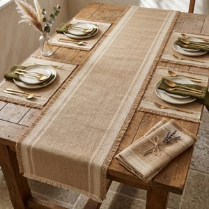Può includere: Un'impostazione rustica per la tavola con un runner da tavolo in iuta beige e tovagliette abbinate. Posate dorate e piatti bianchi sono disposti su tovaglioli verdi. Un vaso con piante secche completa l'arredamento.