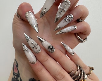 Puede incluir: Uñas largas y puntiagudas, blancas y plateadas. Cada uña presenta un diseño plateado, incluyendo cruces, flor de lis y tachuelas. Las uñas están adornadas con detalles plateados sobre un fondo claro.