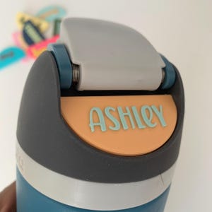 Può includere: Una borraccia turchese e grigia con il nome "ASHLEY" in lettere azzurre su sfondo arancione. La bottiglia ha un coperchio ribaltabile grigio e una fascia argentata. Altri accessori colorati sono visibili sullo sfondo.