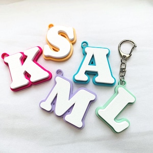 Peut inclure: Plusieurs porte-clés lettres colorés, formant le mot "KAML". Chaque lettre blanche est bordée de rose, jaune, bleu turquoise, violet et vert. Un porte-clés est équipé d'un anneau argenté.
