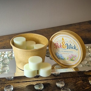 Könnte beinhalten: Eine Sammlung von Pick & Wick Wachsschmelzen. Das Bild zeigt einen runden Behälter mit dem Etikett "Spring Awakening Wax Melts" und einen Kraftpapierbecher, der mit hellgrünen Wachsschmelzen gefüllt ist. Mehrere Wachsschmelzen sind vor dem Becher gestapelt.