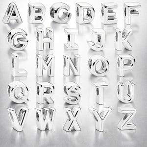Può includere: Una collezione di ventisei perline con lettere dell'alfabeto in metallo color argento disposte in una griglia. Ogni perlina presenta una superficie lucida e riflettente con un foro centrale per l'infilatura, dalla A alla Z.