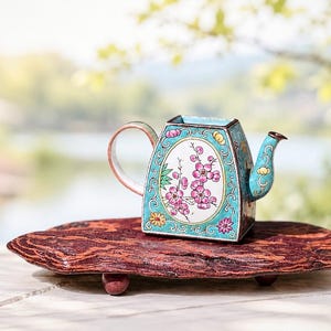 May include: A small, ornate turquoise teapot with a floral design, set on a reddish-brown wooden base. The teapot features a handle, spout, and a decorative oval with pink flowers. The background is blurred, suggesting an outdoor setting.