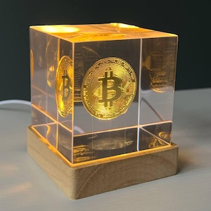 May include: A clear, illuminated cube featuring a golden Bitcoin symbol. The cube is set on a wooden base, with light shining through the Bitcoin design. This decorative piece is a modern interpretation of the cryptocurrency.