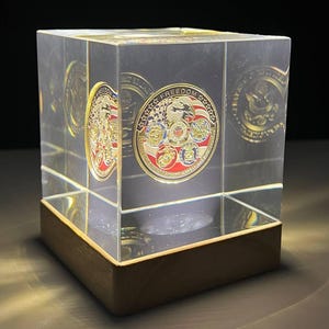 以下が含まれることがあります： 赤、白、青のアクセントが付いた金色のコインを収めた透明なアクリルキューブ。コインには「FREEDOM」の文字と軍事エンブレムが描かれています。キューブは木製の台座の上に置かれています。