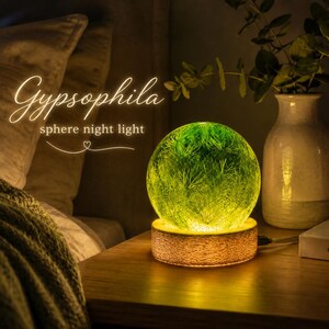 Puede incluir: Una luz nocturna esférica con un diseño de planta Gypsophila verde encapsulado en una esfera transparente, sobre una base circular de madera. El texto "Gypsophila sphere night light" aparece en blanco sobre la lámpara iluminada.