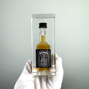 May include: A miniature Jack Daniel's Tennessee Whiskey bottle encased in a clear acrylic block. The bottle has a black label with white text and a black cap. The whiskey inside is amber-colored.