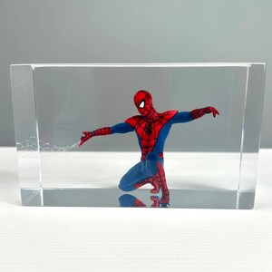 Peut inclure: Figurine de Spider-Man agenouill&eacute;, enferm&eacute;e dans un bloc rectangulaire transparent. Le super-h&eacute;ros porte un costume rouge et bleu, les bras tendus. Le bloc transparent offre une vue claire de la figurine. Le bloc mesure environ 10 cm de haut.