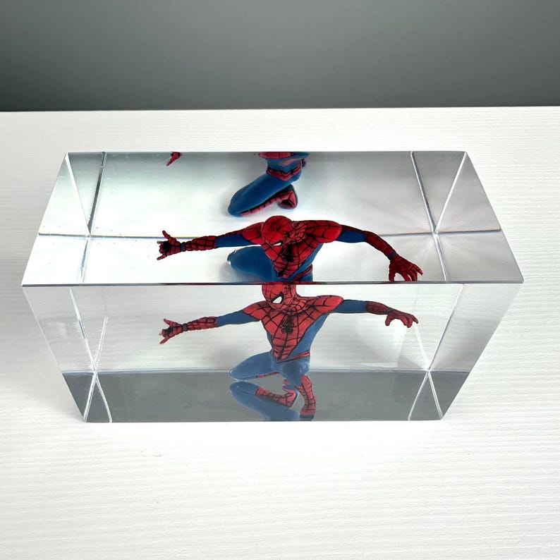 Peut inclure: Prisme rectangulaire transparent avec une figurine de Spiderman &agrave; l'int&eacute;rieur. La figurine, v&ecirc;tue d'un costume rouge et bleu, est en position accroupie. Le prisme refl&egrave;te l'image, cr&eacute;ant un effet miroir. L'arri&egrave;re-plan est gris clair.