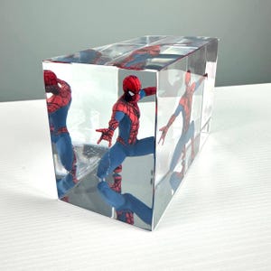 Peut inclure: Un pr&eacute;sentoir transparent en forme de cube avec une figurine de Spider-Man. La figurine est dans une pose dynamique, v&ecirc;tue d'un costume rouge et bleu. Les c&ocirc;t&eacute;s miroit&eacute;s du cube refl&egrave;tent la figurine, cr&eacute;ant plusieurs images.