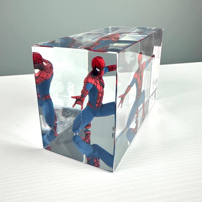 Peut inclure: Un pr&eacute;sentoir transparent en forme de cube avec une figurine de Spider-Man. La figurine est dans une pose dynamique, v&ecirc;tue d'un costume rouge et bleu. Les c&ocirc;t&eacute;s miroit&eacute;s du cube refl&egrave;tent la figurine, cr&eacute;ant plusieurs images.