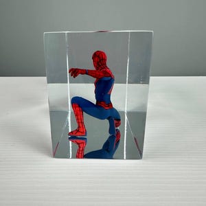 Peut inclure: Figurine de Spider-Man rouge et bleu, enferm&eacute;e dans un bloc rectangulaire transparent. Le super-h&eacute;ros est en position accroupie, avec son reflet visible. L'objet est un objet de collection.