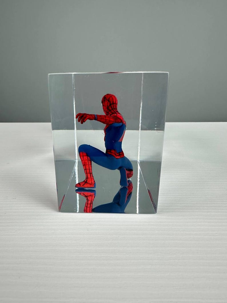 Peut inclure: Figurine de Spider-Man rouge et bleu, enferm&eacute;e dans un bloc rectangulaire transparent. Le super-h&eacute;ros est en position accroupie, avec son reflet visible. L'objet est un objet de collection.